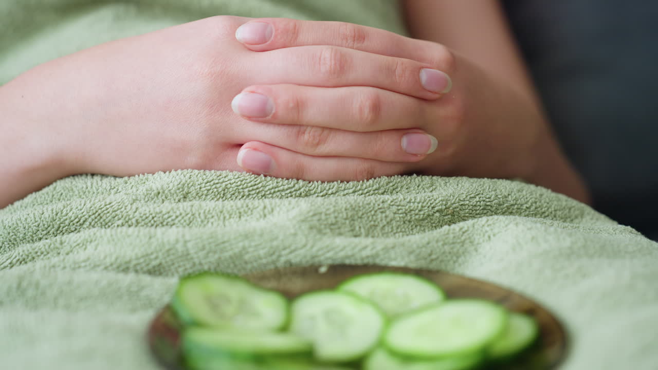 Close-up view of woman's hands folded together, wrapped in towel, with plate of cucumber slices resting on her lap, relaxing skincare routine, wellness, and rejuvenation concept