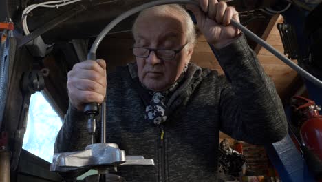 Senior-caucasian-worker-operates-hand-honing-tool-to-shape-a-piece-of-machine,-in-a-small-dark-workshop