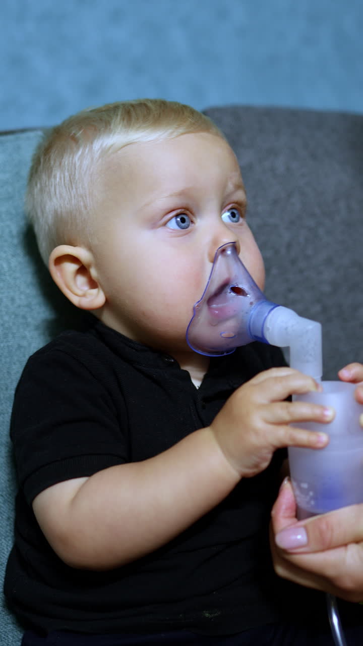 Un bambino piccolo è seduto sul divano e respira con un inalatore. Inalazione terapeutica per bambini. Maschera per inalazione per bambini