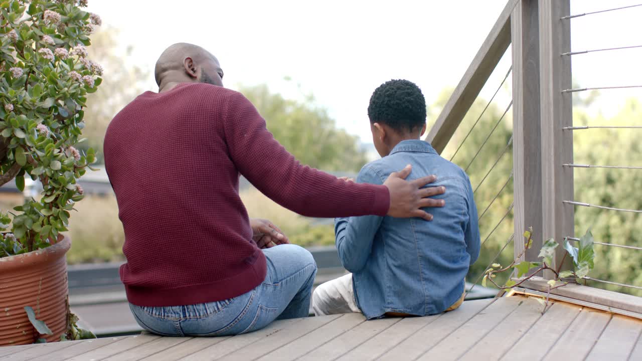 Padre afroamericano e figlio che osservano il giardino sul ponte, rivolti verso il figlio, gli dà una pacca sulla schiena per confortarlo