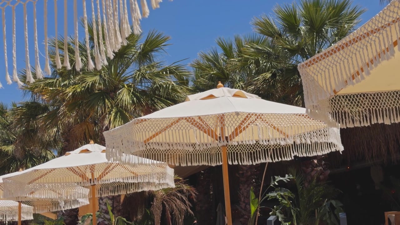 Inquadratura media statica di ombrelloni boho chic con frange che ondeggiano dolcemente con palme tropicali e un cielo azzurro chiaro sullo sfondo in un resort di lusso sulla spiaggia.