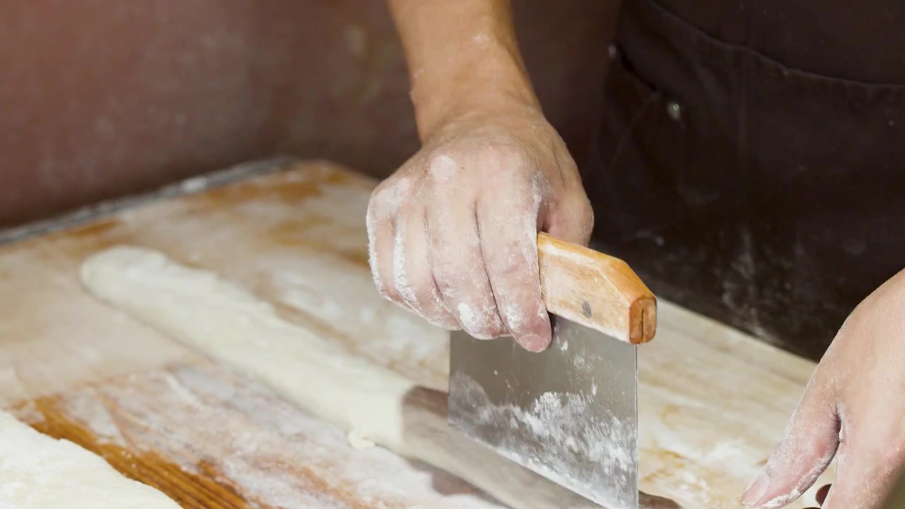 Uno chef usa un raschietto da banco e un mattarello sull'impasto su un tagliere di legno.