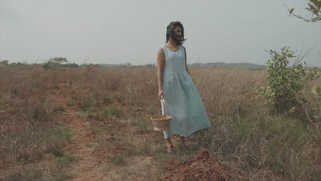 Static-slow-motion-shot-of-a-young-elegant-woman-in-light-blue-dress-with-a-wood-basket-in-her-right-hand-standing-on-a-dry-and-dusty-cornfield