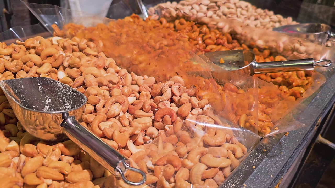 Bulk food store display with roasted cashews piled beside a stainless scoop, plastic bin dividers, golden lighting, and inviting gourmet snack counter for nut and trail mix footage.