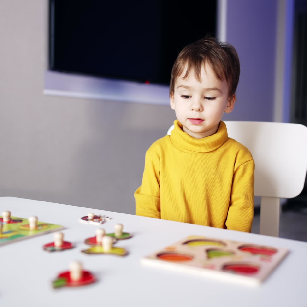 Bambino piccolo carino e intelligente seduto alla scrivania. Il bambino prende due pezzi di puzzle e li unisce. Sfondo sfocato.
