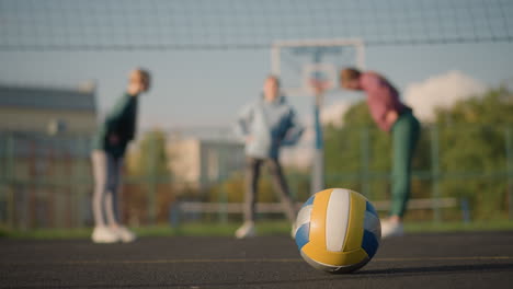 volleyball placed on the ground in an outdoor sport arena with yellow markings visible, athletes in the blurred background engaging in fitness exercises, with focus on volleyball and movement