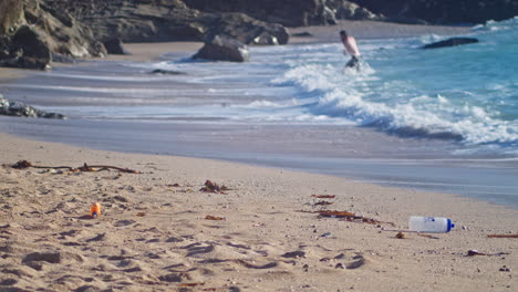 vista al mar de la playa de arena de cornualles con botellas de plástico desechadas y hombre nadando en el fondo- estático