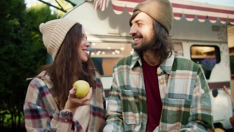 primer plano: una chica morena con una manzana amarilla en la mano y comunicándose con su hombre moreno con una camisa a cuadros verde sentado cerca de su remolque en un campamento durante un picnic fuera de la ciudad en verano