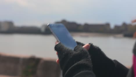 Toma-De-Primer-Plano-De-Una-Mujer-Caminando-Mientras-Escribe-En-Su-Teléfono-Inteligente-Cerca-De-Un-Río-Con-La-Ciudad-Al-Fondo