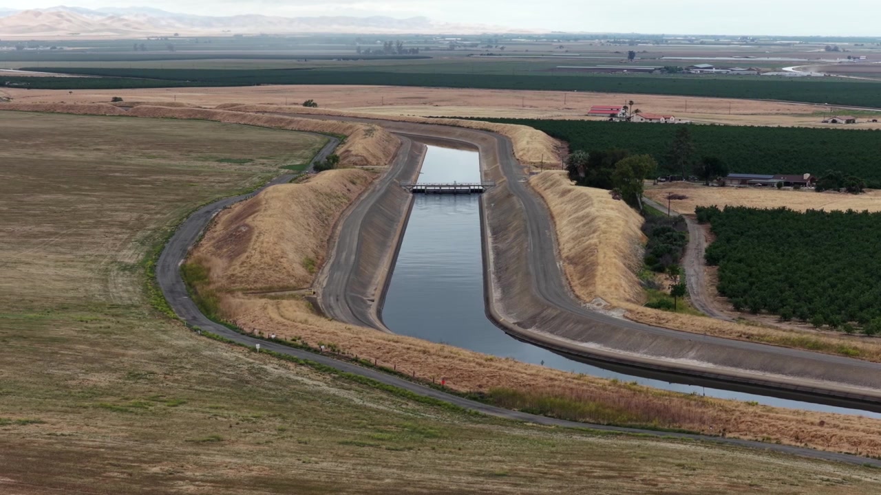 Canale dell'acquedotto che si snoda attraverso una prateria dorata e secca accanto a frutteti verdi, ponte che attraversa a metà con una valle pianeggiante e montagne sfocate visibili in lontananza. California, USA. Ampia ripresa aerea.
