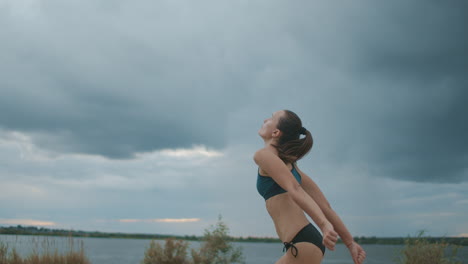 Joven-Deportista-Vestida-Con-Bikini-Está-Jugando-Voleibol-De-Playa-De-Tiro-Medio-Contra-El-Pintoresco-Cielo-Nublado