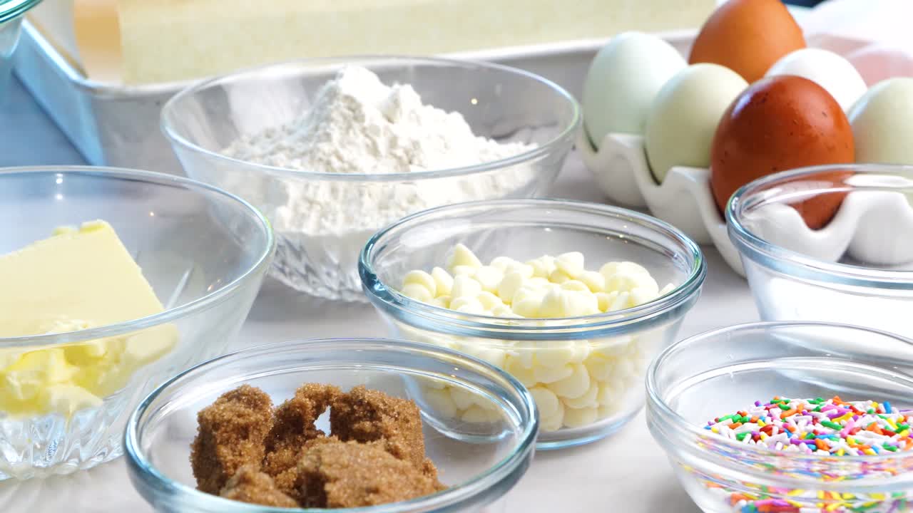 Baking confetti sugar cookies and panning across all the cookie ingredients.. In the kitchen making a dessert. Making confetti cookies with a white background, pink napkin, chocolate chips and eggs