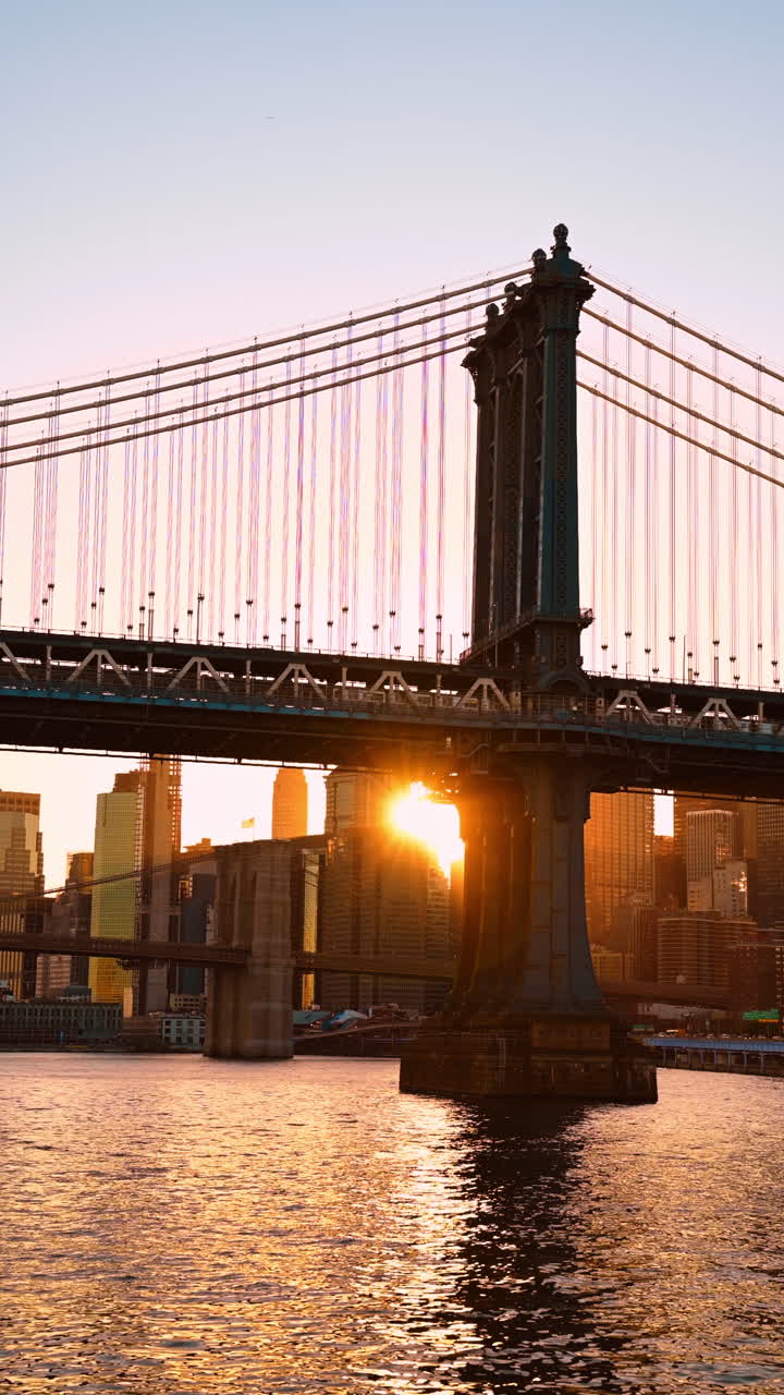 Tramonto tra gli edifici e il Manhattan Bridge. Treno in movimento vicino al ponte. Angolazione dal basso. New York, USA. Video verticale