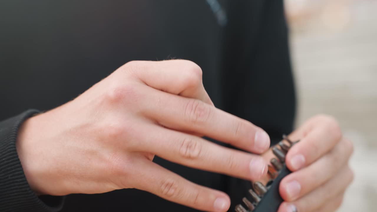 Tampilan dekat tangan putih memegang mata obeng, memilih kepala obeng kecil dengan ujung jari yang mantap, bagian logam dan perangkat ringkas di luar ruangan, keterampilan manual yang presisi dan konsentrasi taktil, intim