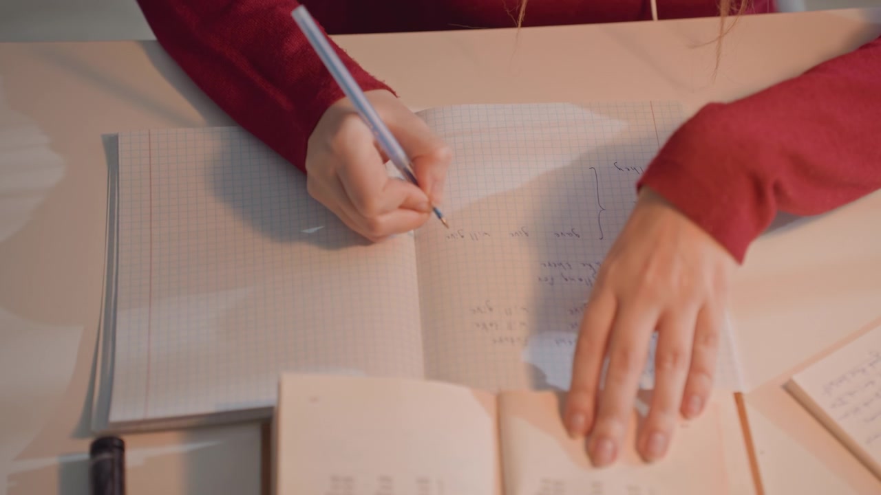 Young woman in red long sleeve garment writes mathematical notes. Scene includes ink bottle like object near materials. Light colored table holds various study items