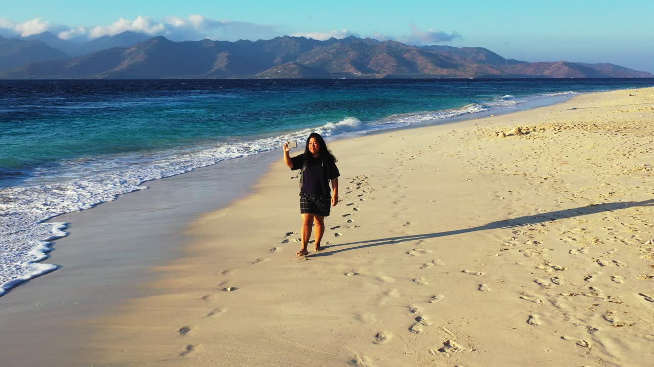 A young woman taking photos on her camera at a tropical beach and splashing in the small waves crashing on the shore