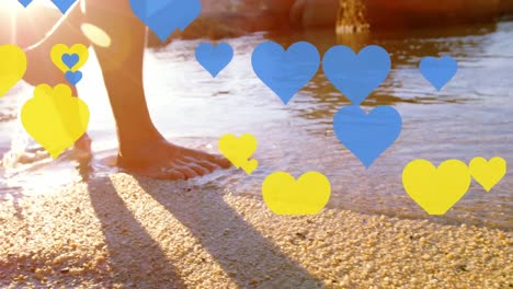 Multiple-blue-and-yellow-heart-icons-floating-against-low-section-of-man-walking-on-the-beach