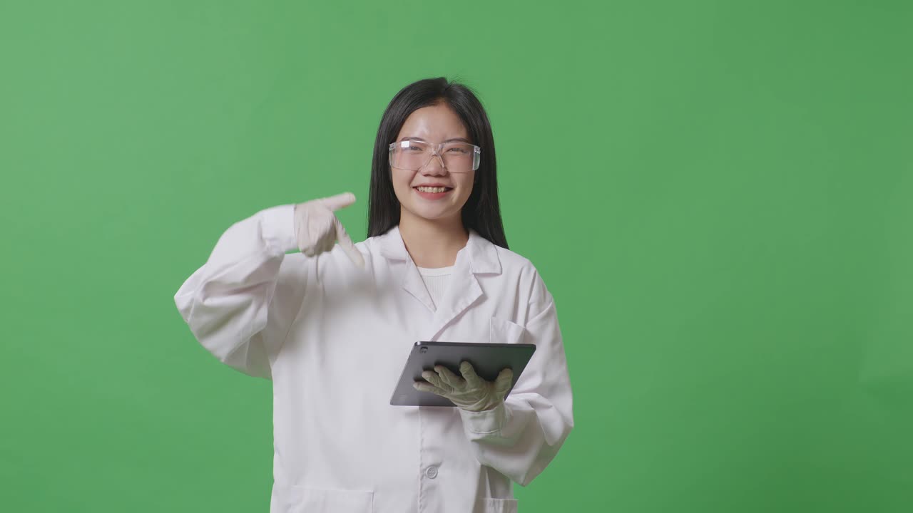아시아 여성 과학자가 미소 짓고 실험실의 녹색 화면 배경에 서서 태블릿을 가리키고 있습니다.