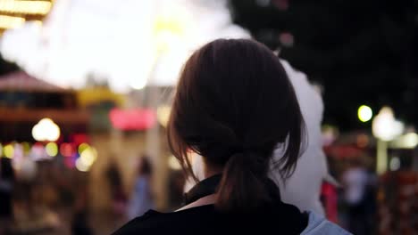 Backside-view-of-a-girl-walking-in-amusement-park,-taking-a-cotton-candy.-Short-haired-blonde-woman-wearing-transparent-glasses