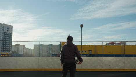 man standing on urban sports field with soccer ball in hand, looking around, surrounded by a backdrop of city buildings, bright yellow walls, and a clear blue sky illuminated by sunlight