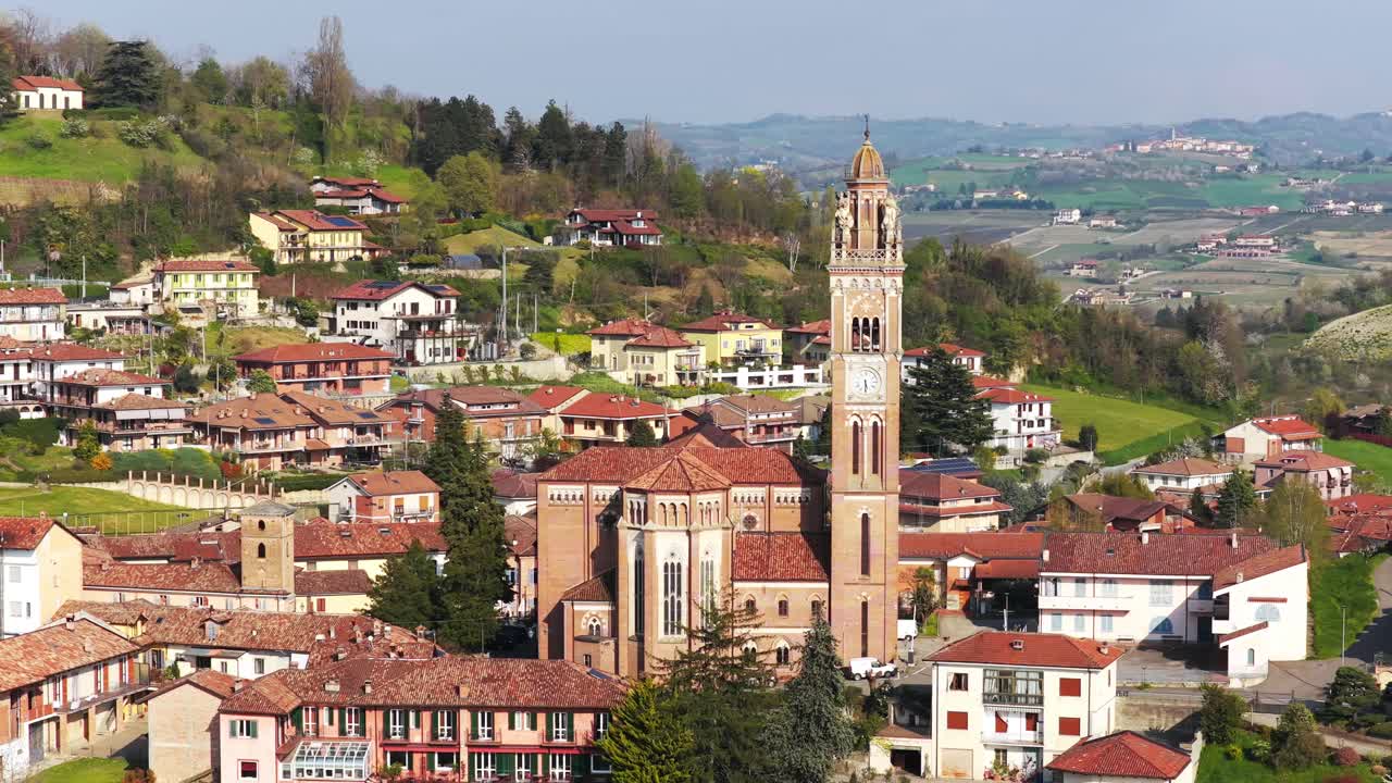 Orbiting aerial shot of Monforte d’Alba, showcasing its iconic church tower, charming red-roofed houses, and rolling hills of the Langhe countryside under clear spring skies