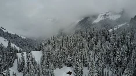 Drone-flying-over-remote-cabin-in-woods-in-winter-nestled-on-cliff-edge