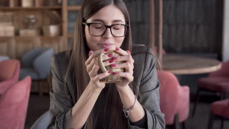 beautiful young woman in a glasses is sitting in a cafe, holding a cup of coffee and sipping