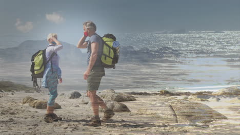 happy caucasian senior couple wearing facemasks hiking by the coast over fast moving clouds