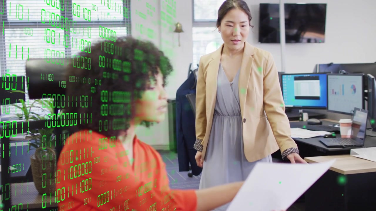 Seated woman turning paper to colleague, both leaning in, reviewing tech charts under green binary