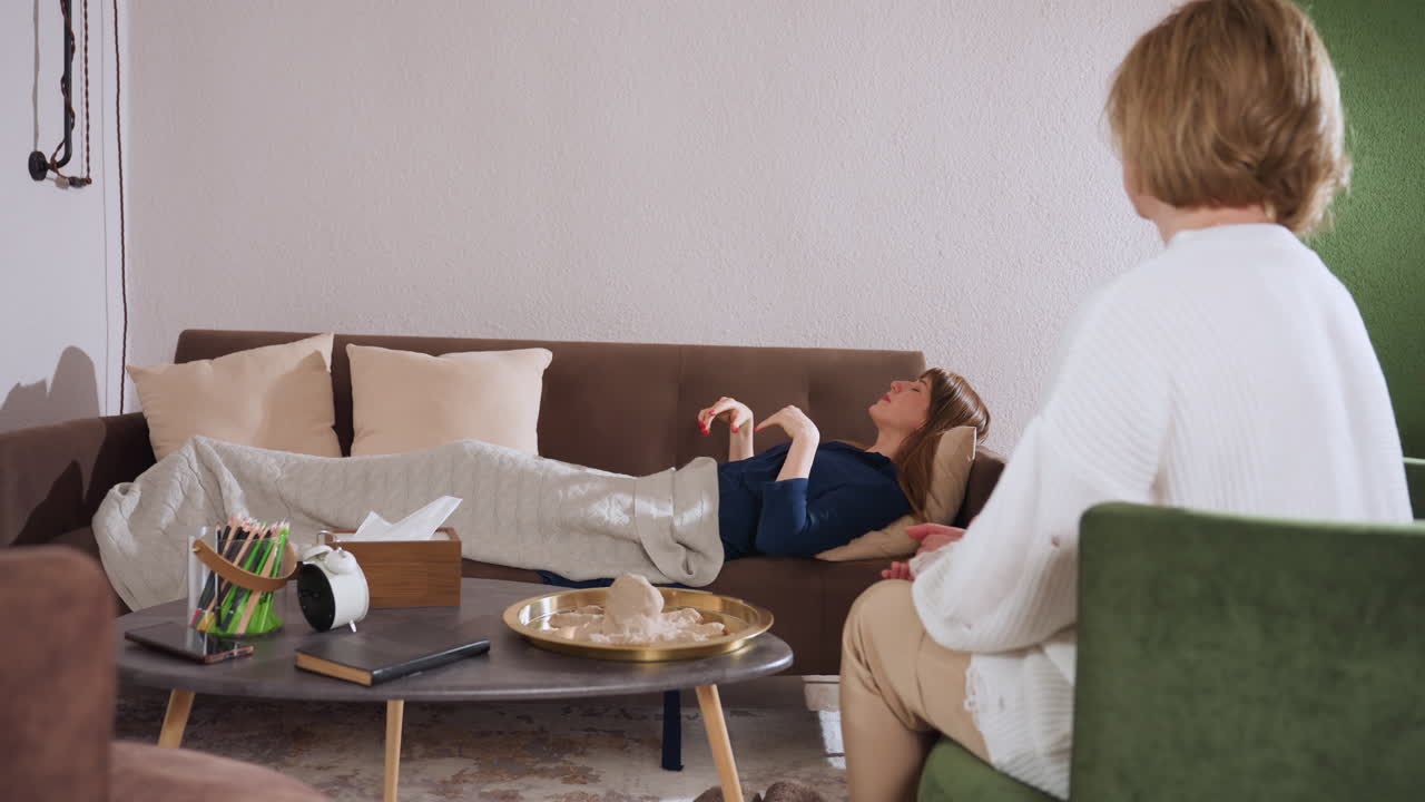 Lady in blue gown relaxes on couch during therapy session, pulling soft blanket over herself while specialist observes nearby in peaceful setting with cozy decor and warm ambient lighting