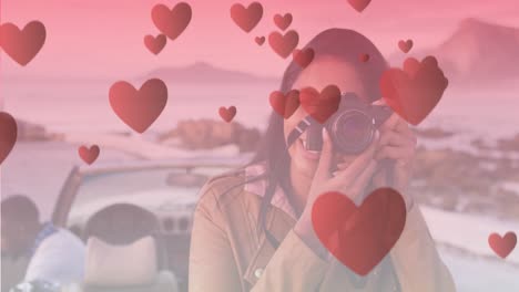 Animation-of-hearts-floating-over-happy-biracial-woman-taking-photo-at-cabriolet