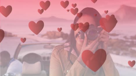 animation of hearts floating over happy biracial woman taking photo at cabriolet