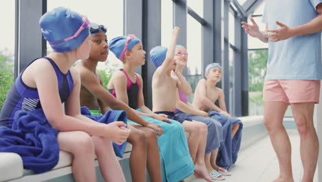 male coach giving children in swimming class briefing as they sit on edge of indoor pool