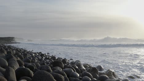 hazy ocean shore view in iceland with waves spraying mist, bright sunshine, rounded stones on beach