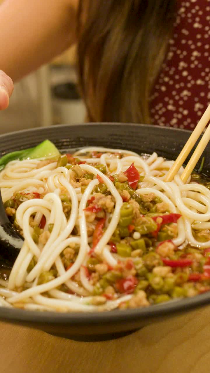 Mulher mexe sopa de noodles picante com colher preta e pauzinhos em um ambiente de restaurante com iluminação acolhedora.