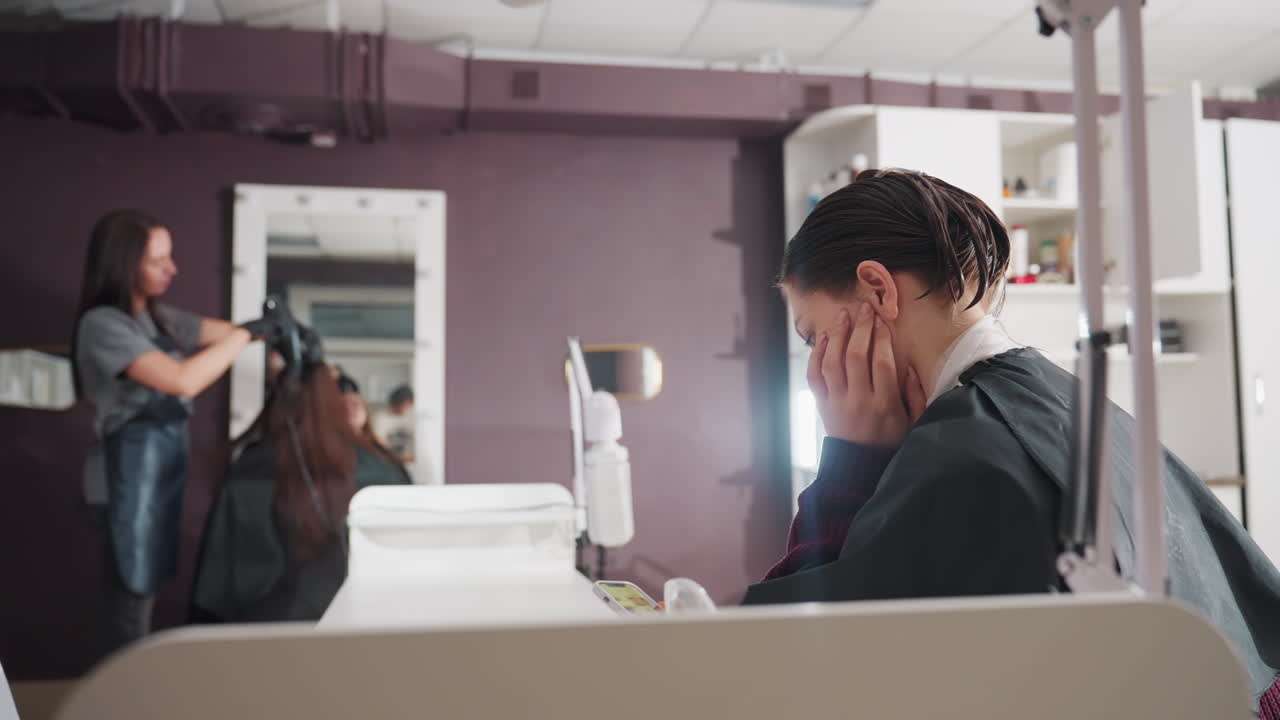 스타일리스트가 거울 앞에서 작업하는 동안 얼굴을 가리고 있는 고객, 배경에는 고객에게 집중하는 스타일리스트, 전경에는 불안한 자세를 보이는 백인, 선반과 부드러운 조명이 있는 아담한 살롱 인테리어