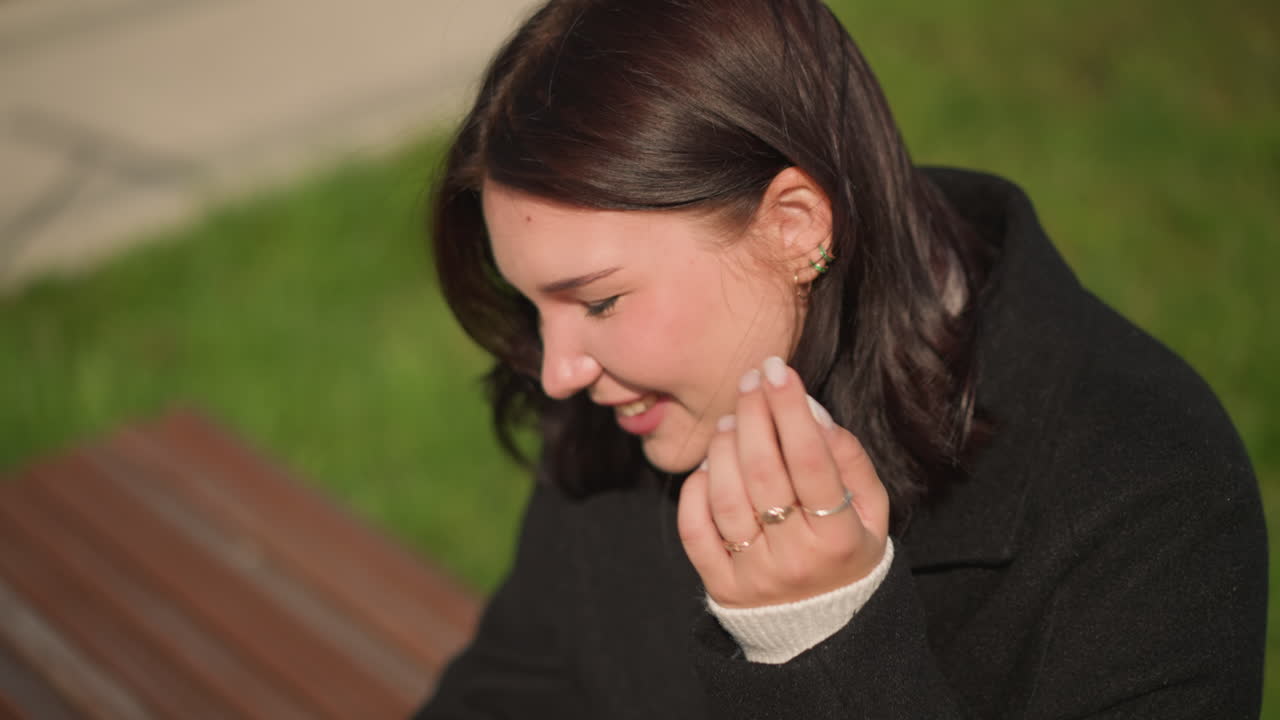 공립 공원에서 머리카락을 조정하는 동안 웃는 검은 코트를 입은 여성의 클로즈업, 귀걸이가 보이고, 손가락이 우아한 반지로 장식되어, 배경에 부드러운 초록색으로 밝은 야외 설정
