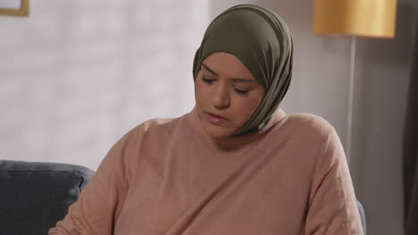 muslim woman wearing hijab sitting on sofa at home reading or studying the quran 1