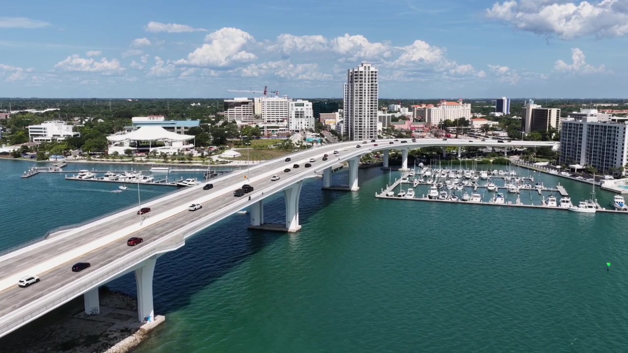 Un ponte a terrapieno si inarca su un porto turchese con auto che attraversano verso lo skyline del centro, un porto turistico fiancheggiato da barche e torri sul lungomare nella zona della baia di Clearwater, Florida. Ripresa aerea con drone in avvicinamento.
