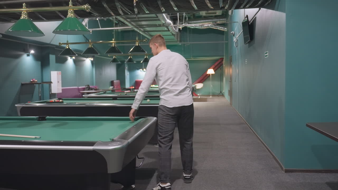 Back shot of player in white shirt and grey trousers walking into billiard room, touching green pool table. Modern sports club setting with overhead lamps, stylish interior atmosphere