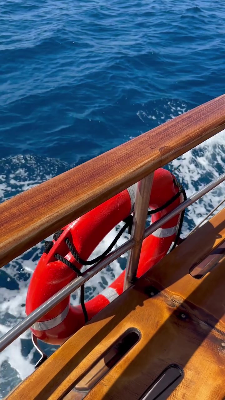 Video per social media che mostra il ponte di una barca con salvagente rosso, ringhiera in legno e scia spumeggiante sopra le acque blu intenso della caldera, durante il viaggio verso Nea Kameni, Santorini, Grecia