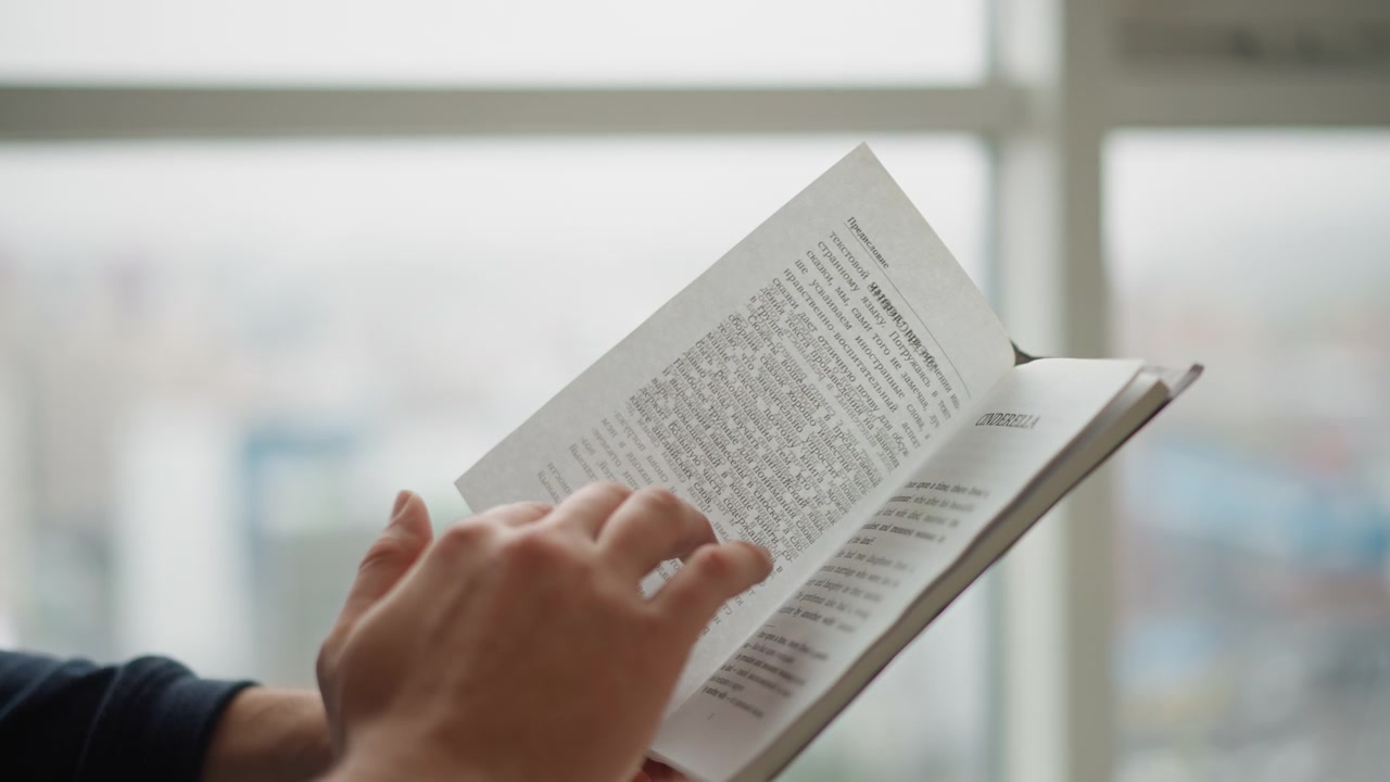 Close up of hands turning pages in open book with text and images visible soft natural light through window background reading paper closeup lighting indoor