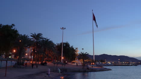 Eine-Flagge-Jordaniens-An-Einem-Sandstrand-Weht-Im-Wind-In-Der-Stadt-Aqaba-Im-Hintergrund-Von-Palmen