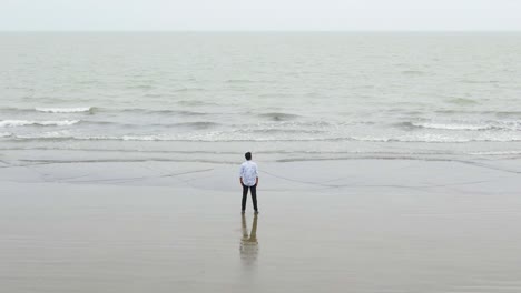 Toma-Inversa-De-Un-Dron-De-Un-Hombre-Parado-Solo-Junto-Al-Mar-Y-Mirando-Triste-O-Melancólicamente-El-Horizonte-Gris-En-Un-Día-Nublado,-Mar-De-Kuakata,-Bangladesh