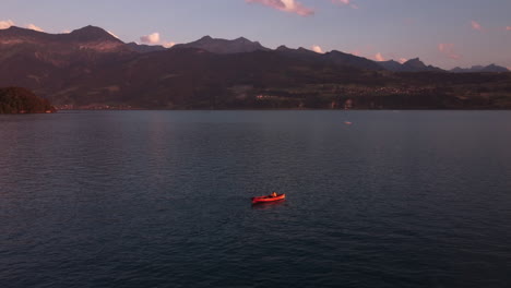 Top-down-aerial-view-of-a-people-paddling-in-a-red-paddling-boat-on-the-deep-turquoise-alpine-lake-Thunersee-in-Switzerland-during-sunset