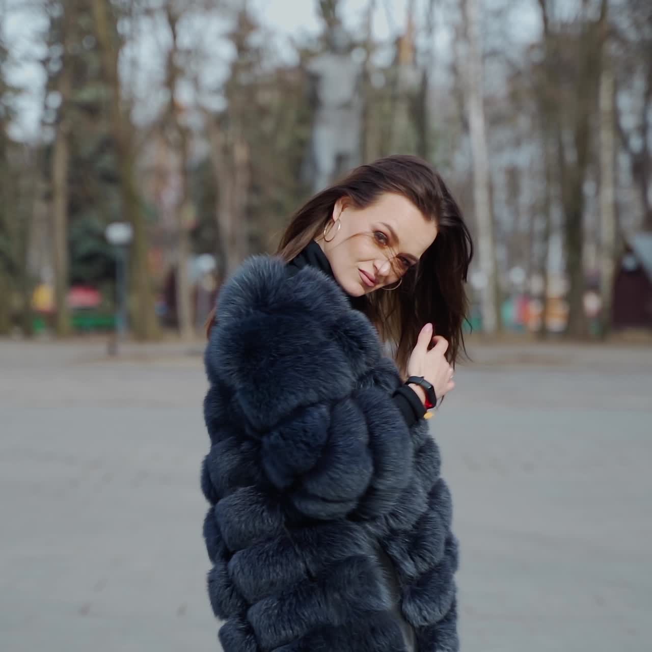 Pretty model in fur coat. Beautiful young woman is spinning happily in the park. Charming girl with long dark hair posing on camera outside.
