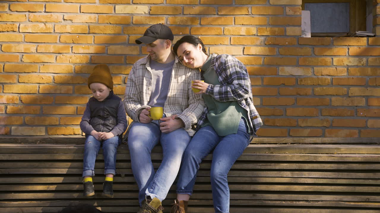 Valkoihoinen pariskunta puhumassa poikansa kanssa heidän juodessaan teetä maalaistalon pihalla. Heidän koiransa makaa maassa heidän lähellään.