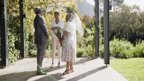 mujer afroamericana oficiando la ceremonia de boda de una pareja biracial mayor en el jardín, cámara lenta