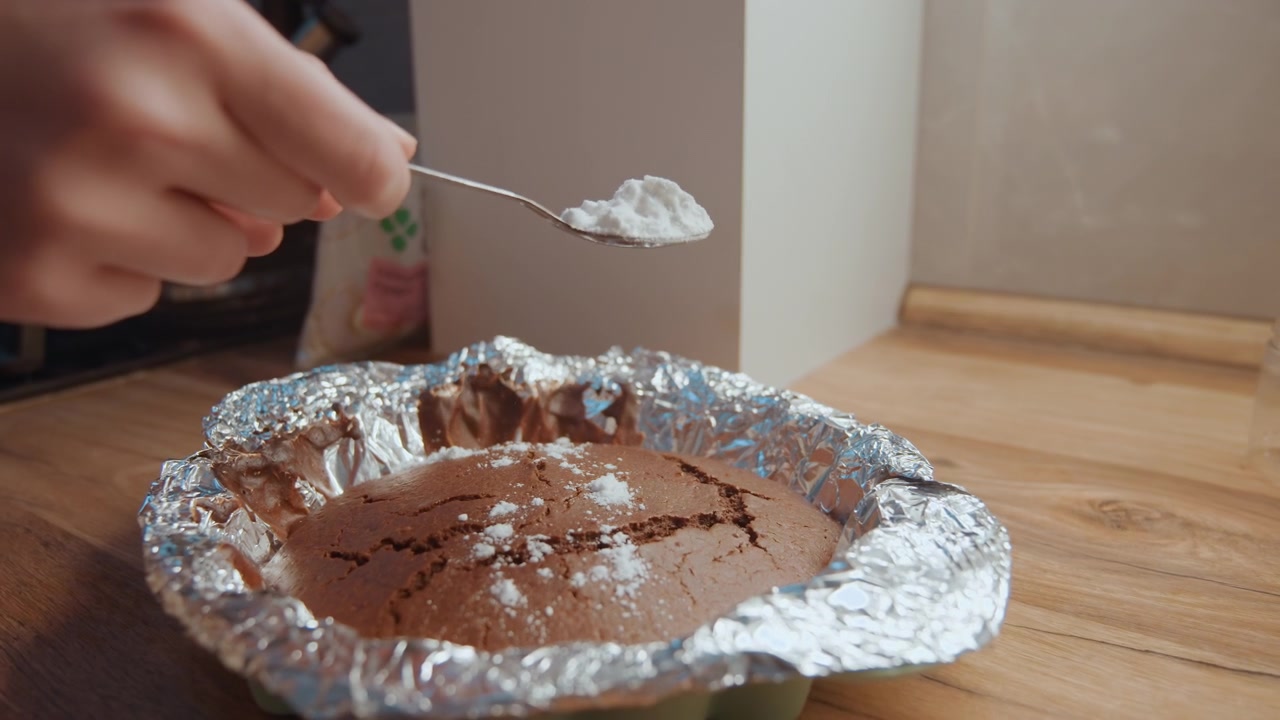 Primer plano de tarta de chocolate siendo espolvoreada con azúcar glas por una mano firme sobre un molde forrado con papel de aluminio en una encimera de madera, luz cálida de cocina, panadero casero terminando el postre, movimiento de cuchara creando delicados detalles