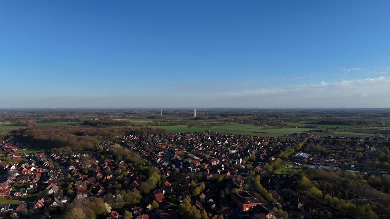 American small town with housing area and blossom trees in spring season. Aerial forward wide shot. Blue sky and sunlight with rotating wind turbines in distance. Suburb neighborhood in United States